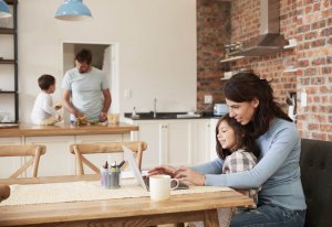 Family of four in a modern kitchen; the mother is working on her laptop with her child in her lap; the father is working in the kitchen and talking to the little boy sitting on the counter