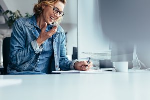 Woman working in an office, talking on a mobile phone, and writing down notes in a notepad