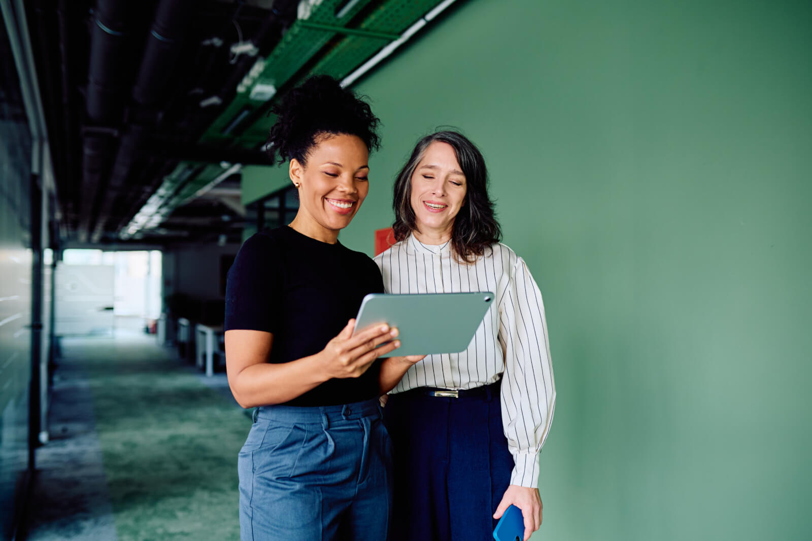 Two smiling colleagues looking at a tablet together in an office hallway